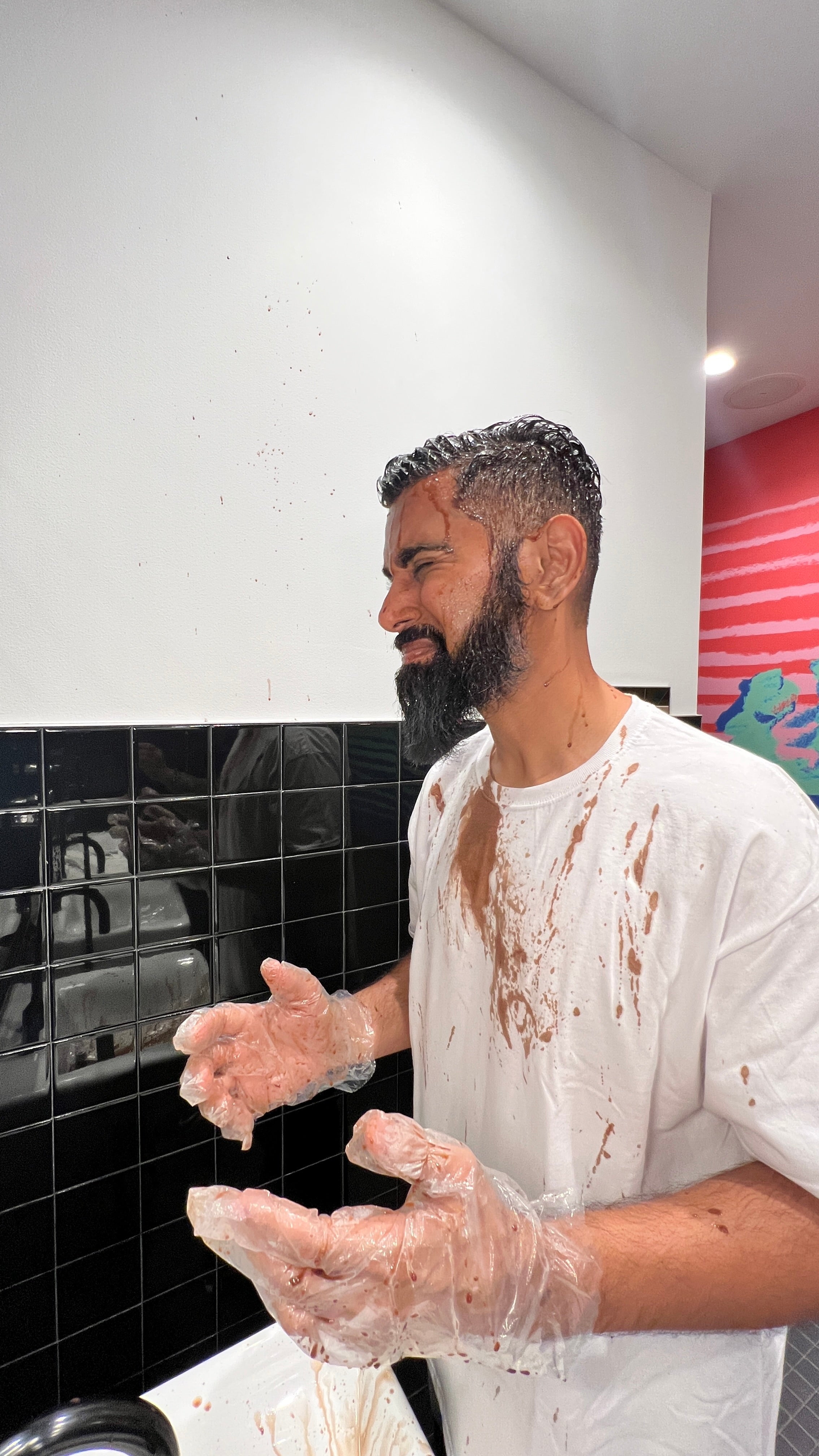 A man with dye dripping onto his face and white shirt, holding up his stained disposable gloves.
