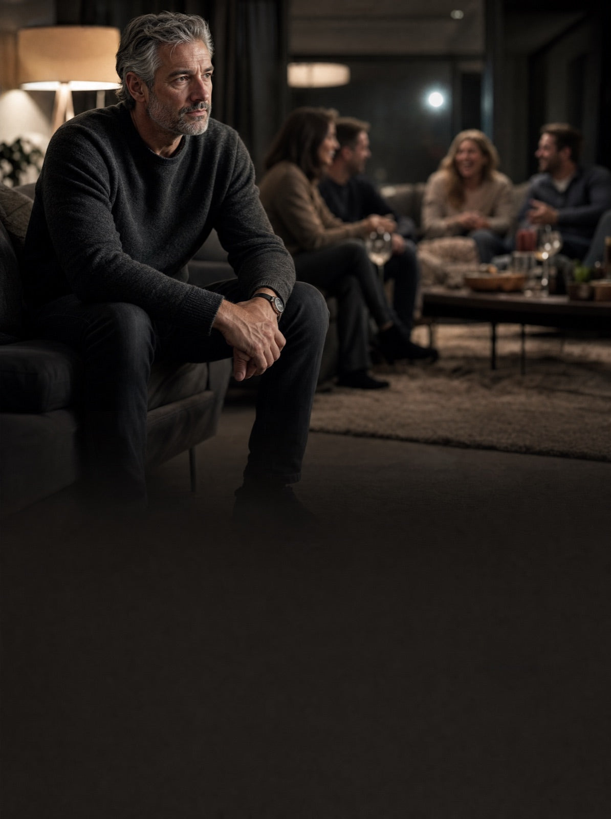 A gray-haired man sitting apart from friends at an evening gathering.