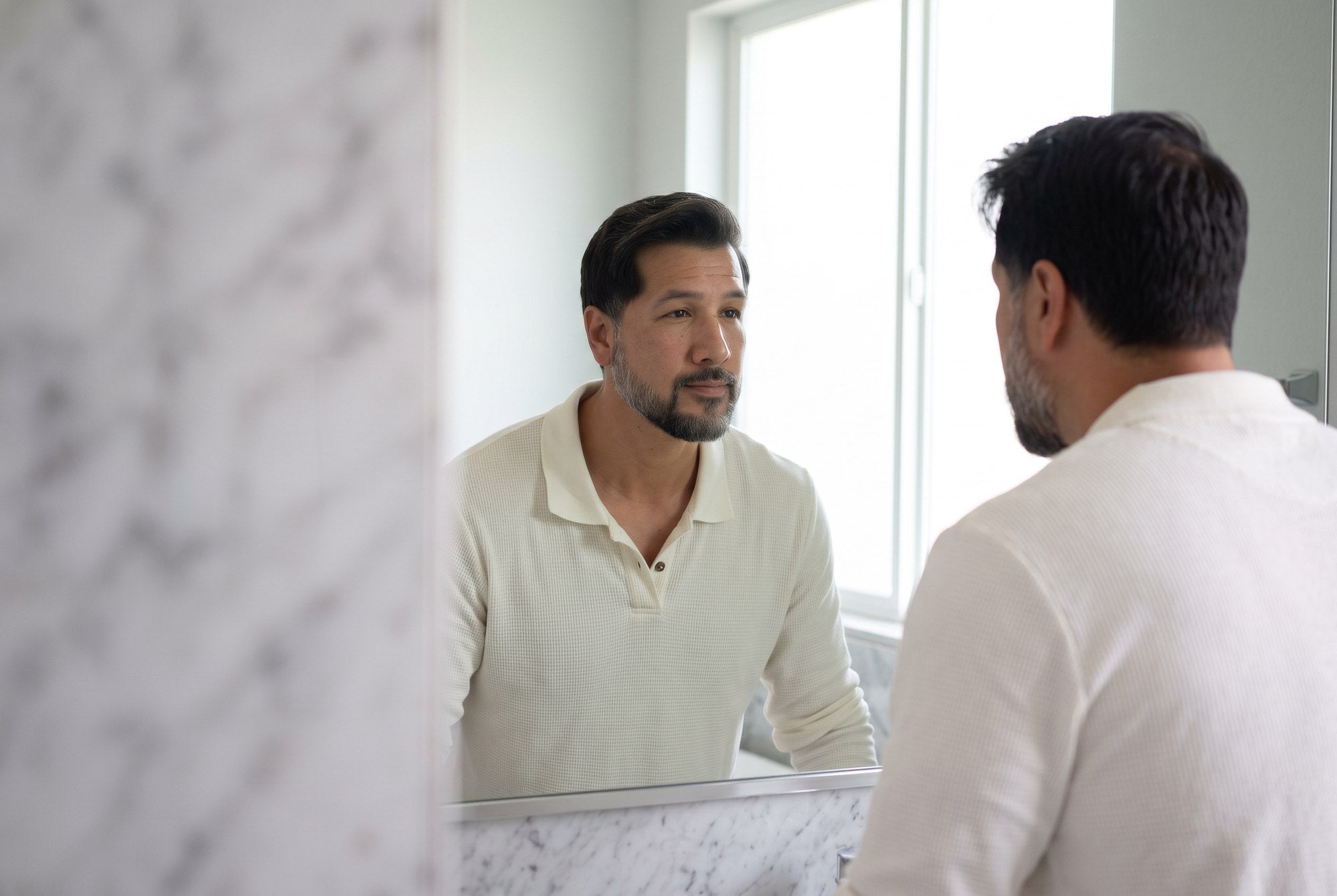 A man with gray hair looking thoughtfully at himself in a bathroom mirror.
